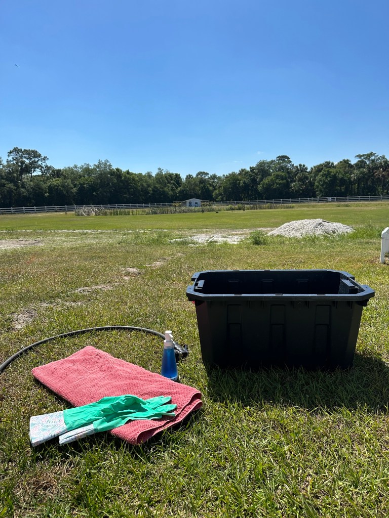 outside on the green grass is a black storage bin, a red towel, hose, blue soap and green gloves.